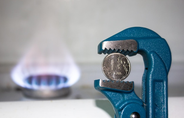 Синий трубный ключ, сжимающий монету номиналом 1 рубль, на фоне зажженной газовой горелки.